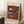 May include: Framed abstract art poster with orange and teal geometric shapes including striped triangles, circles, and angular forms in Kandinsky's modernist style. Light wood frame leans against white wall corner next to wooden side table with white coffee cup and saucer. Cream textured rug and beige fabric visible at bottom.
