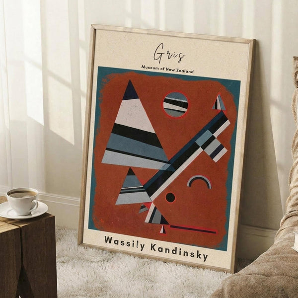 May include: Framed abstract art poster with orange and teal geometric shapes including striped triangles, circles, and angular forms in Kandinsky's modernist style. Light wood frame leans against white wall corner next to wooden side table with white coffee cup and saucer. Cream textured rug and beige fabric visible at bottom.