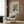 May include: Van Gogh sunflowers museum poster displayed on wood-paneled wall above beige upholstered armchair with geometric patterned pillow. Brass floor lamp with white circular base stands nearby next to dark round side table with open book. Warm natural lighting filters through white curtains, highlighting the poster's yellow sunflowers in green vase against blue background. Modern minimalist reading nook with wooden flooring.