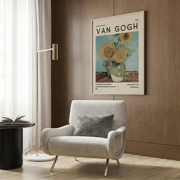 May include: Van Gogh sunflowers museum poster displayed on wood-paneled wall above beige upholstered armchair with geometric patterned pillow. Brass floor lamp with white circular base stands nearby next to dark round side table with open book. Warm natural lighting filters through white curtains, highlighting the poster's yellow sunflowers in green vase against blue background. Modern minimalist reading nook with wooden flooring.