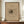 May include: A framed abstract sketch print displayed on a wooden credenza against white wainscoted walls. The artwork features dark expressive lines and gestural marks on beige paper, mounted in a gold frame. A small white ceramic vase and stack of books sit nearby on the warm wood surface.