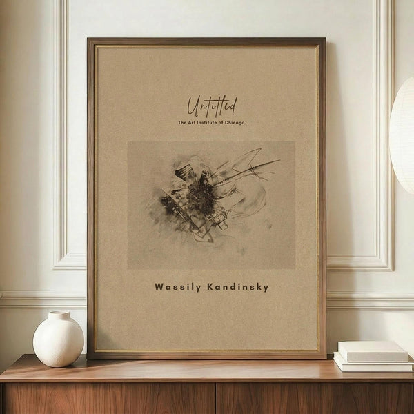 May include: A framed abstract sketch print displayed on a wooden credenza against white wainscoted walls. The artwork features dark expressive lines and gestural marks on beige paper, mounted in a gold frame. A small white ceramic vase and stack of books sit nearby on the warm wood surface.