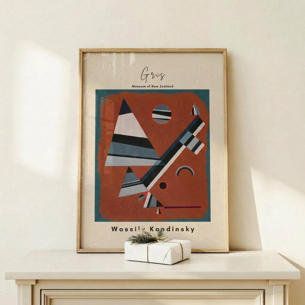 May include: Framed abstract art print in wooden frame leaning against white wall on light wood surface. The artwork features geometric shapes including striped triangles, circles, and angular forms in rust orange, teal, black, white, and gray colors. Museum exhibition poster format with "Wassily Kandinsky" text below the artwork. Small wrapped gift box with twine sits in front of the frame.