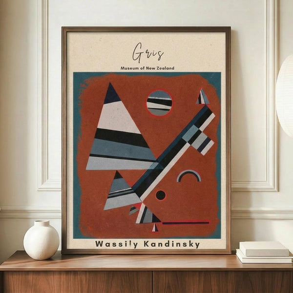 May include: Framed abstract geometric art print in warm orange and teal tones, featuring triangular and linear shapes in Kandinsky's distinctive style. The poster shows "Gris" at top with "Museum of New Zealand" and "Wassily Kandinsky" text below the artwork. Displayed on wooden sideboard alongside white ceramic vase and stacked books in modern interior with cream walls and white trim.