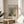 May include: Framed Van Gogh museum poster featuring his Three Sunflowers painting displayed on a round wooden side table next to a beige upholstered chair. The poster shows yellow sunflowers in a green vase against turquoise background, with museum-style text and cream matting in thin gold frame. Soft natural lighting creates shadows on white wall, with plant leaves visible in corner.