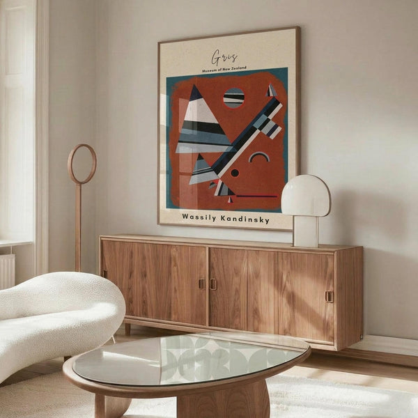 May include: Abstract geometric art poster with red-orange background featuring triangular and linear shapes in black, white, and blue tones, displayed in thin wooden frame above mid-century walnut credenza. Modern living room setting with cream textured chair, glass oval coffee table, and minimalist white table lamp creating warm, contemporary interior design aesthetic.