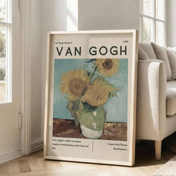 May include: Framed museum-style poster featuring Van Gogh's sunflower painting with yellow blooms in green vase against turquoise background. Light wood frame leans against white wall next to beige upholstered chair in bright, naturally-lit room with large windows and hardwood floors.