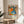 May include: Vibrant exhibition poster with colorful polka dots, flowers, and organic shapes in orange, blue, green, and pink tones. The framed artwork sits on a light wood round stool beside a beige upholstered chair. Natural sunlight creates soft shadows across white walls and sheer curtains, with a green plant visible in the corner of this minimalist room setting.