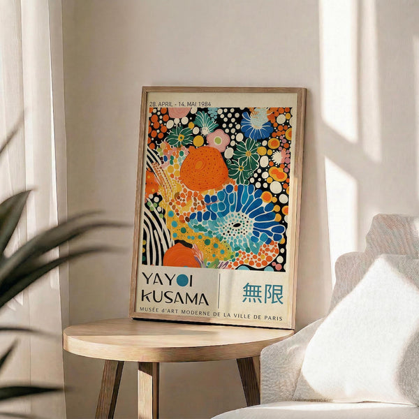 May include: Vibrant exhibition poster with colorful polka dots, flowers, and organic shapes in orange, blue, green, and pink tones. The framed artwork sits on a light wood round stool beside a beige upholstered chair. Natural sunlight creates soft shadows across white walls and sheer curtains, with a green plant visible in the corner of this minimalist room setting.