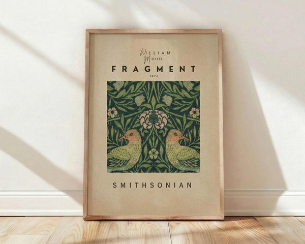 May include: Framed vintage botanical poster featuring William Morris design with two green and peach birds surrounded by ornate foliage and flowers on dark green background. Light wooden frame leaning against white wall on natural wood floor with soft window lighting casting shadows.
