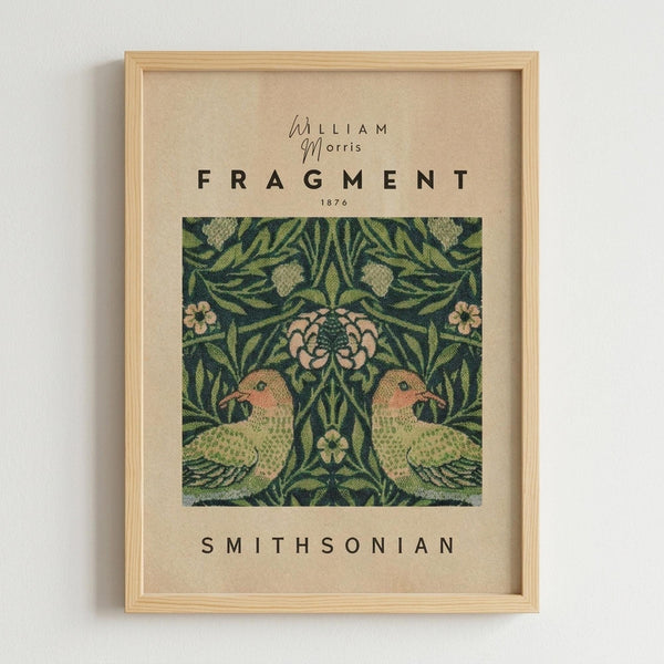 Framed art print featuring a William Morris textile design from 1876 with two green parrots facing each other surrounded by ornate foliage, pink flowers, and decorative botanical motifs on a dark green background. The vintage-style poster has cream colored text reading "William Morris," "FRAGMENT," "1876," and "SMITHSONIAN" on a beige background. Displayed in a light wood frame against a white wall.