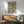 May include: Framed botanical art print with green background and cream-colored flowing acanthus leaves and flowers in ornate Victorian style, displayed above wooden mid-century modern sideboard with table lamp, next to sage green upholstered armchair with wooden arms in bright naturally-lit room with white walls and hardwood floors.