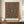 May include: Framed exhibition poster featuring William Morris's intricate Holland Park carpet design with ornate floral patterns in deep teal, rust red, and golden tones. The botanical motifs include stylized flowers and curving vines arranged in symmetrical medallions. Displayed on a wooden sideboard alongside a cream ceramic vase and stacked books against white wainscoted walls.