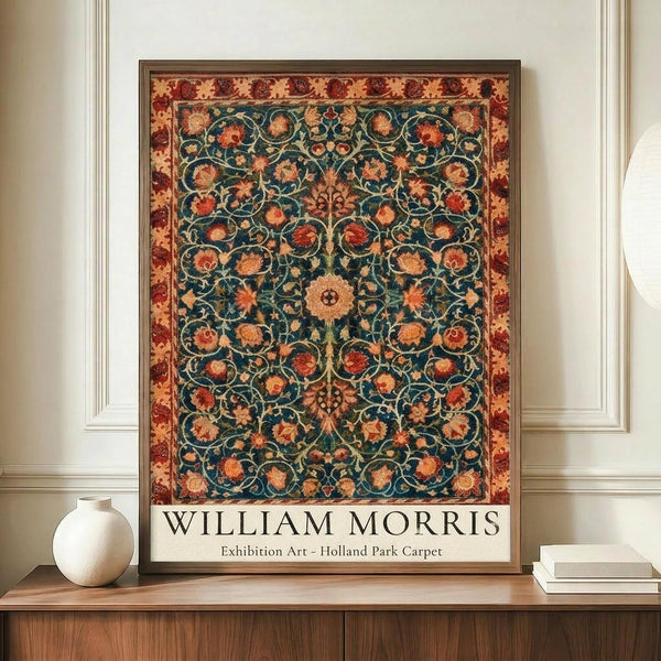 May include: Framed exhibition poster featuring William Morris's intricate Holland Park carpet design with ornate floral patterns in deep teal, rust red, and golden tones. The botanical motifs include stylized flowers and curving vines arranged in symmetrical medallions. Displayed on a wooden sideboard alongside a cream ceramic vase and stacked books against white wainscoted walls.