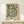 May include: Framed botanical art print with green background and cream-colored ornate floral scrollwork pattern displayed on light wood console table. Small white wrapped gift with green sprig sits beside the golden wood-framed poster against neutral beige wall.