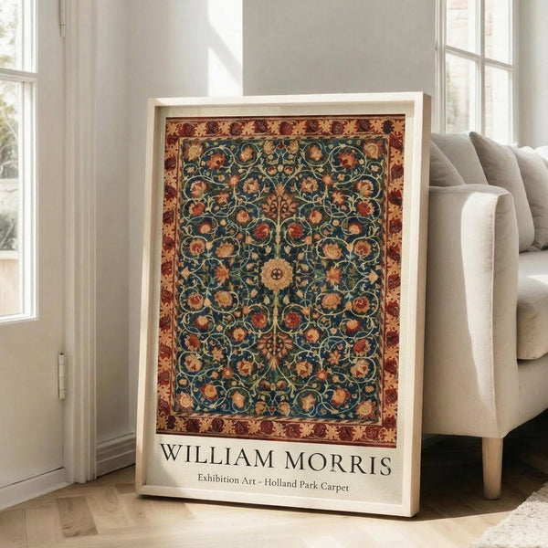 May include: Framed exhibition poster featuring William Morris's ornate carpet design with intricate floral patterns in deep blues, warm oranges, and burgundy tones. White matted frame leans against white wall next to beige upholstered sofa in bright, sunlit living room with tall windows and light wooden floors.