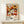 May include: Framed vintage-style exhibition poster featuring colorful daisy and sunflower pattern in warm orange, yellow, white and brown tones. Shows dates from 1984, artist name, and Japanese characters. Light wood frame sits on pale wooden surface next to small wrapped gift with twine and greenery sprig against cream wall.