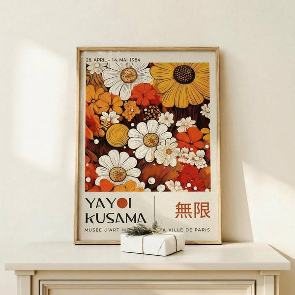 May include: Framed vintage-style exhibition poster featuring colorful daisy and sunflower pattern in warm orange, yellow, white and brown tones. Shows dates from 1984, artist name, and Japanese characters. Light wood frame sits on pale wooden surface next to small wrapped gift with twine and greenery sprig against cream wall.