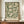 May include: Framed poster with sage green background featuring cream-colored ornate floral scrollwork and leaves in William Morris style, displayed on wooden sideboard next to white ceramic vase and stacked books in elegant room with white wainscoting and cream walls