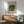May include: Framed William Morris botanical poster with green pattern featuring two birds among stylized leaves and flowers, displayed above a wooden sideboard with white ceramic lamp. Mid-century modern sage green upholstered chair with wooden arms sits nearby. Warm natural lighting creates soft shadows on white wall in minimalist room setting.