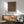 May include: Framed William Morris exhibition art print hanging on white wall above wooden sideboard with table lamp, shown next to sage green mid-century armchair and round wooden coffee table. The poster features an ornate carpet design with intricate floral patterns in deep teal, orange, and burgundy colors with decorative border, displayed in natural lighting.