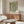 May include: Framed botanical poster with green and cream floral pattern displayed above wooden sideboard in modern living room. White sculptural chair and glass-top coffee table visible. Poster shows flowing leaves and flowers in vintage Arts and Crafts style with text at bottom. Natural lighting creates shadows across warm wood furniture and neutral walls.