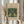 May include: Hands holding a framed botanical art print featuring two symmetrical green birds surrounded by ornate foliage and flowers in William Morris' signature Arts and Crafts style. The print displays rich emerald greens with coral pink floral accents on cream paper within a natural wood frame. Dried pampas grass stems are held alongside, and the scene is set against soft white curtains in natural lighting, creating an elegant home decor styling.