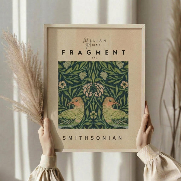 May include: Hands holding a framed botanical art print featuring two symmetrical green birds surrounded by ornate foliage and flowers in William Morris' signature Arts and Crafts style. The print displays rich emerald greens with coral pink floral accents on cream paper within a natural wood frame. Dried pampas grass stems are held alongside, and the scene is set against soft white curtains in natural lighting, creating an elegant home decor styling.