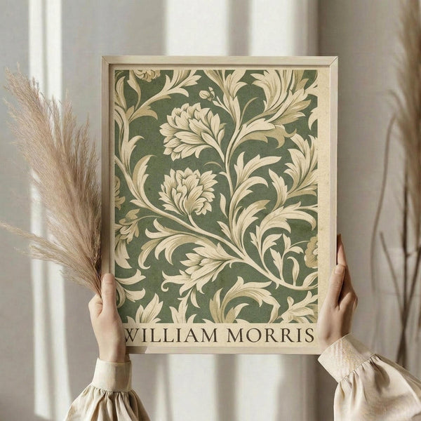 May include: Hands holding a framed botanical print featuring sage green background with cream-colored ornate floral scrollwork pattern in William Morris style. Light wood frame, person wearing beige ruffle-sleeved top, holding dried pampas grass stems. Soft natural lighting against white curtained window background creates elegant vintage aesthetic.