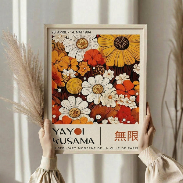 May include: Hands holding a framed vintage exhibition poster featuring colorful retro flowers in orange, yellow, white and red tones against a dark background. The artwork shows daisies, sunflowers and abstract blooms with dotted centers. Light wood frame against soft neutral curtains with dried pampas grass visible to the left. The poster displays museum exhibition text and Japanese characters at the bottom.