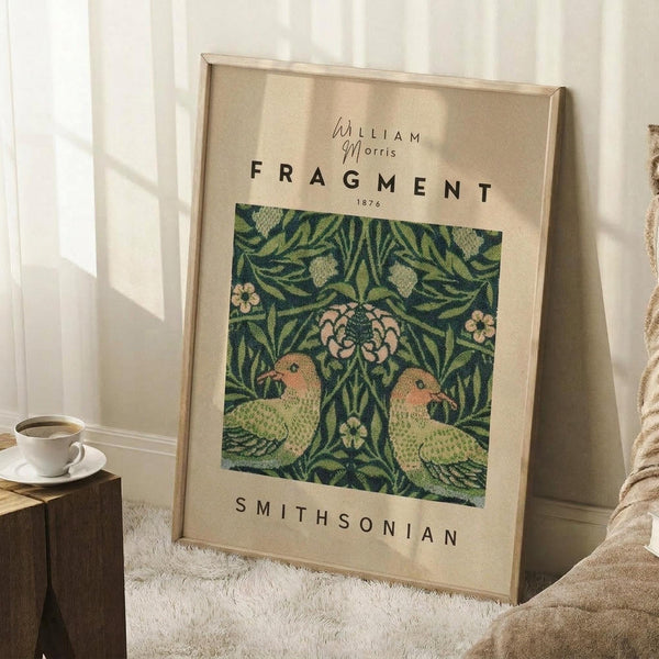 May include: Framed William Morris botanical art print featuring two green parrots amid leafy vines and pink flowers on dark background. Light wood frame leaning against white wall corner next to wooden side table with white coffee cup and saucer. Cream colored blanket visible on white textured rug.