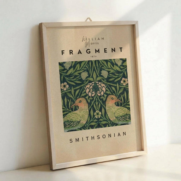 May include: Framed art print leaning against white wall showing William Morris textile design fragment from 1876. Features two green parrots surrounded by ornate foliage and pink flowers on dark background. Cream matting with "FRAGMENT" and "SMITHSONIAN" text. Light wood frame with visible hanging hardware. Natural lighting creates soft shadows on wall.