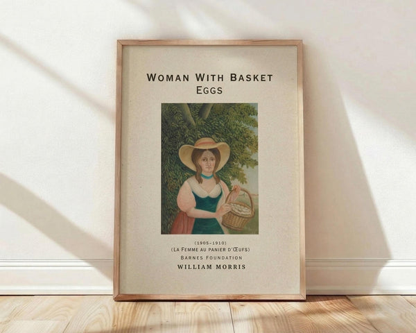 May include: Framed vintage art print showing a woman in period dress with wide-brimmed hat holding a wicker basket of eggs, standing beneath green foliage. The print features muted earth tones and classical painting style, displayed in a natural wood frame with cream matting. The frame leans against a white wall on light wooden flooring with soft natural lighting creating gentle shadows.