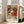 May include: Framed botanical art print leaning against white wall beside beige upholstered sofa in bright living room. Print features ornate orange and red chrysanthemum flowers with teal and green decorative leaves in Victorian Arts and Crafts style. Natural wood frame, approximately 2-3 feet tall. Sunlight streams through tall windows onto light wooden floors with partial view of area rug.
