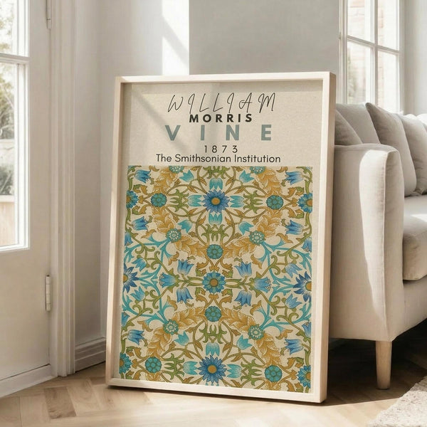 May include: Framed botanical art print featuring William Morris floral vine pattern in blue, green and gold tones, displayed in light wood frame leaning against white wall next to beige upholstered chair in bright, airy living room setting with hardwood floors.