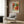 May include: Framed botanical art print featuring vibrant orange and coral flowers with green foliage in vintage William Morris style, displayed on wood paneled wall above beige upholstered armchair with geometric patterned pillow, brass floor lamp with white base nearby, creating warm modern interior scene with natural lighting from window.