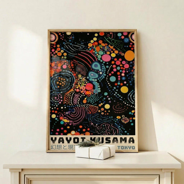 May include: Framed poster with vibrant dot painting on black background featuring swirling patterns in rainbow colors - pink, orange, blue, yellow, green circles and curved lines. Text reads "YAYOI KUSAMA" and "TOKYO" at bottom. Wooden frame sits on light wooden surface next to small white gift box with dried plant sprig against cream wall.