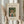May include: Hands holding a framed art print featuring a woman in a wide-brimmed hat and green dress carrying a wicker basket of eggs, surrounded by lush green foliage. The print has cream matting with black text identifying it as a William Morris piece from 1905-1910. One hand also holds dried pampas grass stems against a soft, neutral background with window light.