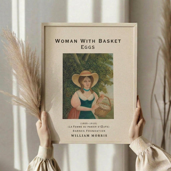 May include: Hands holding a framed art print featuring a woman in a wide-brimmed hat and green dress carrying a wicker basket of eggs, surrounded by lush green foliage. The print has cream matting with black text identifying it as a William Morris piece from 1905-1910. One hand also holds dried pampas grass stems against a soft, neutral background with window light.