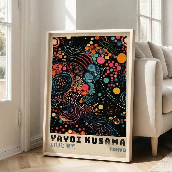 May include: Framed art print with colorful dot pattern on black background featuring swirling circles, spirals, and geometric shapes in vibrant blues, oranges, pinks, and yellows. Text reads "YAYOI KUSAMA" with Japanese characters and "TOKYO" below. Light wood frame leaning against white wall next to beige sofa in bright room with large windows.