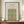 May include: A framed vintage botanical poster featuring an intricate blue, gold, and green floral vine pattern displayed on a wooden sideboard. The cream-colored print shows ornate flowering vines with detailed leaves and blooms in a symmetrical design. A small white ceramic vase and stacked books sit nearby on the warm wood surface against cream wainscoted walls.