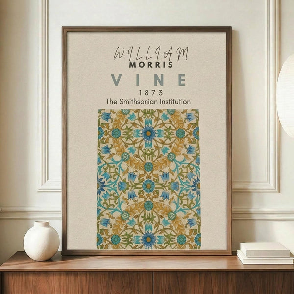May include: A framed vintage botanical poster featuring an intricate blue, gold, and green floral vine pattern displayed on a wooden sideboard. The cream-colored print shows ornate flowering vines with detailed leaves and blooms in a symmetrical design. A small white ceramic vase and stacked books sit nearby on the warm wood surface against cream wainscoted walls.