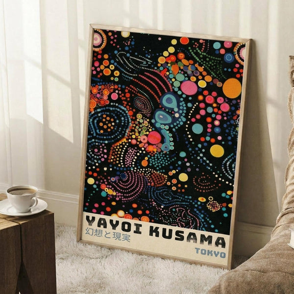 May include: Framed art print leaning against white wall corner featuring colorful dot painting on black background with vibrant circles, spirals and organic patterns in rainbow hues. Japanese text at bottom reads Yayoi Kusama with Tokyo below. Natural wood frame, displayed near white coffee cup on wooden block and beige knit throw on white textured rug.