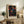 May include: Framed art print with colorful abstract dot pattern in blues, oranges, pinks, and yellows on black background, displayed on rustic wooden sideboard next to fern plant, books, ceramic vase and lamp in neutral-toned living room setting with beige armchair visible.