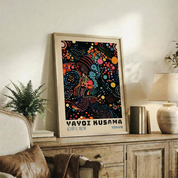 May include: Framed art print with colorful abstract dot pattern in blues, oranges, pinks, and yellows on black background, displayed on rustic wooden sideboard next to fern plant, books, ceramic vase and lamp in neutral-toned living room setting with beige armchair visible.