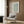 May include: Framed botanical print with sage green willow leaf pattern displayed above a beige upholstered armchair with geometric patterned pillow. Modern brass floor lamp beside chair, dark round side table with open book. Warm wood paneled wall backdrop with natural lighting from sheer curtained window.