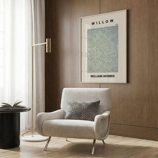 May include: Framed botanical print with sage green willow leaf pattern displayed above a beige upholstered armchair with geometric patterned pillow. Modern brass floor lamp beside chair, dark round side table with open book. Warm wood paneled wall backdrop with natural lighting from sheer curtained window.