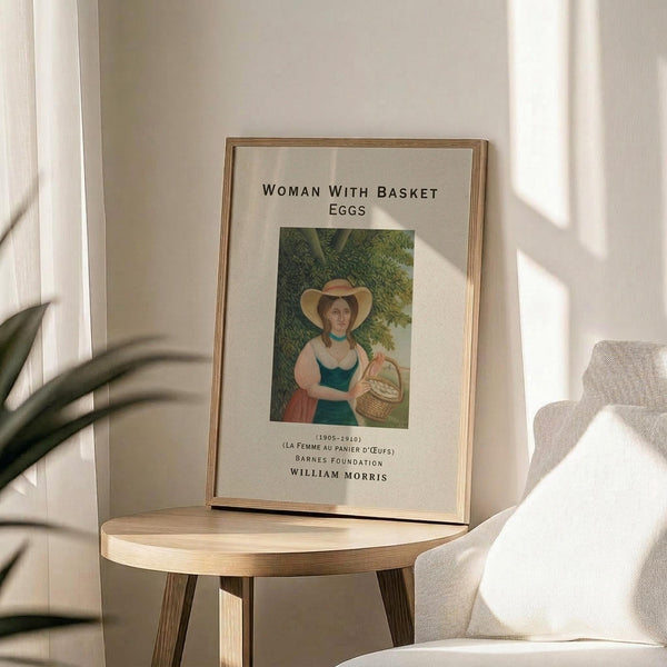 May include: Framed art print featuring a woman in a wide-brimmed hat and teal dress holding a wicker basket, set against green foliage background. The poster has white matting with black text reading "Woman with Basket Eggs" and attribution details. Displayed on a round wooden side table next to a beige upholstered chair, with a potted plant visible and natural sunlight casting shadows on white walls.