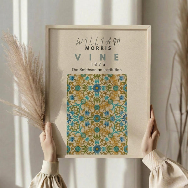 May include: Hands holding a framed poster featuring William Morris's Vine pattern from 1873 in blues, golds, and greens with intricate floral motifs. The beige matted frame is displayed against white curtains with dried pampas grass visible to the left. Person wears a cream-colored blouse with gathered sleeves.