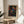 May include: Colorful abstract art print in wooden frame featuring vibrant dots, circles, and swirling patterns on black background in blues, reds, oranges, and yellows. Frame sits on round wooden side table next to beige upholstered chair with plant leaves visible. Soft natural lighting creates shadows on neutral wall. Japanese text visible below artwork. Contemporary home decor styling.